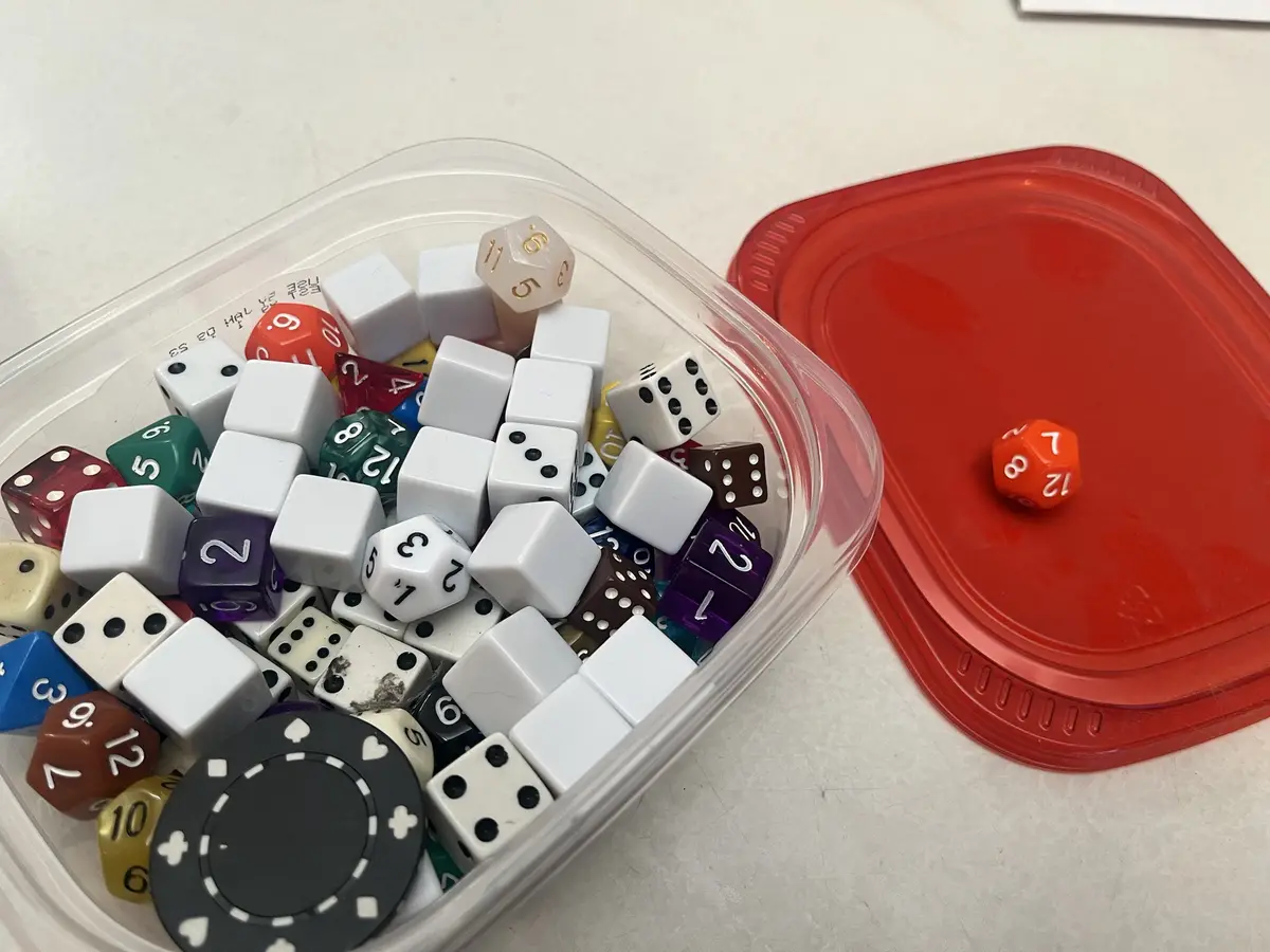 A clear plastic container filled with various colorful polyhedral dice and one black poker chip sits on a white surface…