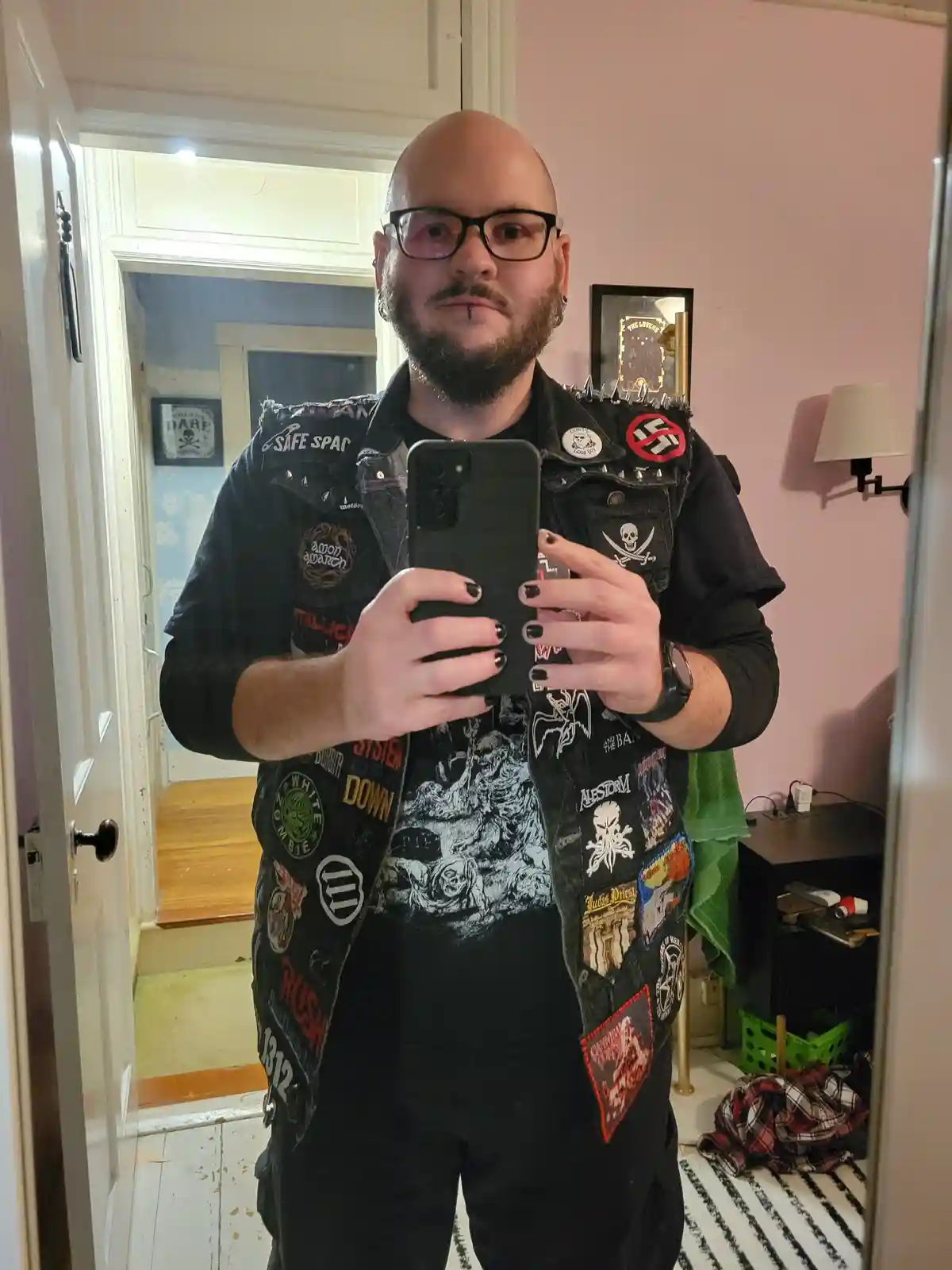 A bald man with glasses and a beard takes a mirror selfie while wearing a heavily patched black denim vest adorned with…