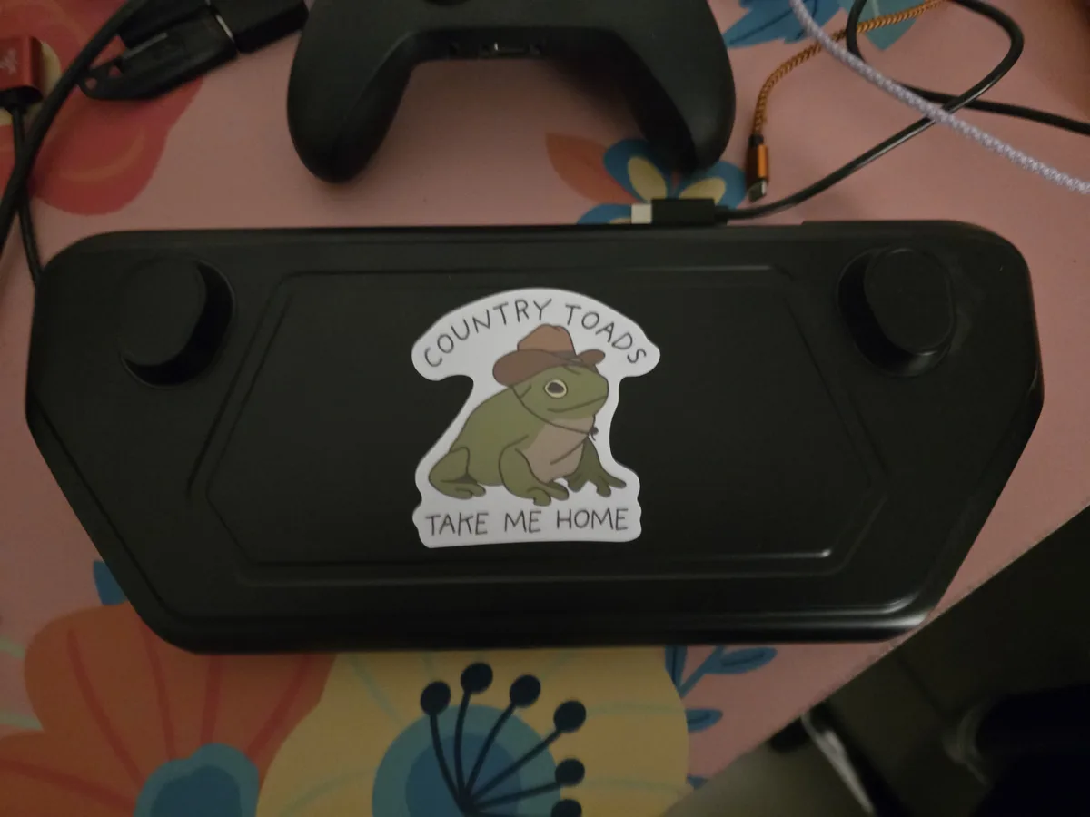 A black handheld gaming device rests on a floral-patterned surface, featuring a sticker of a green toad wearing a cowboy…