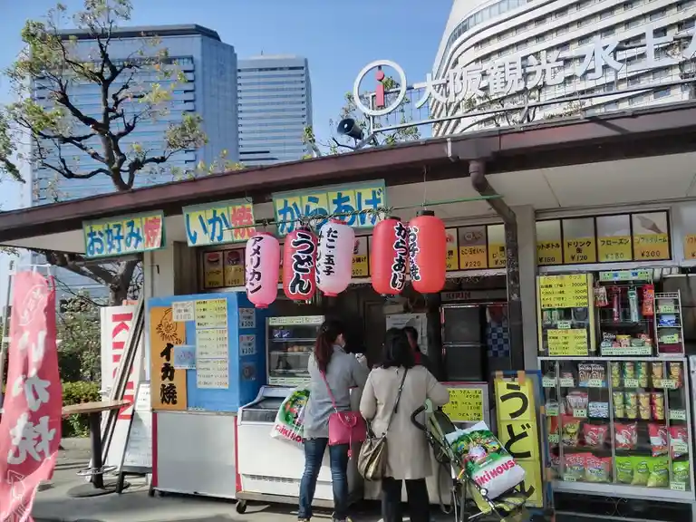 A street food stall in Japan with customers ordering from a counter, surrounded by colorful signs and red lanterns with…
