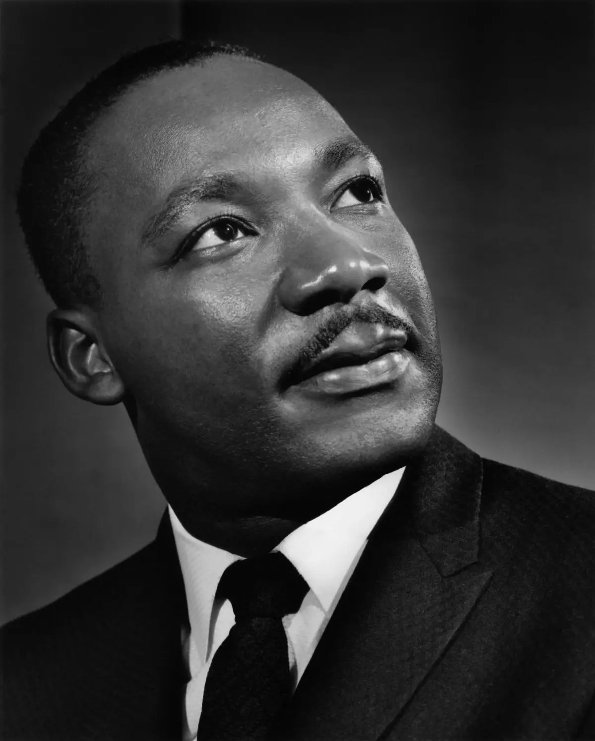 A black-and-white portrait of Martin Luther King Jr. looking upward with a thoughtful expression, wearing a suit and tie…