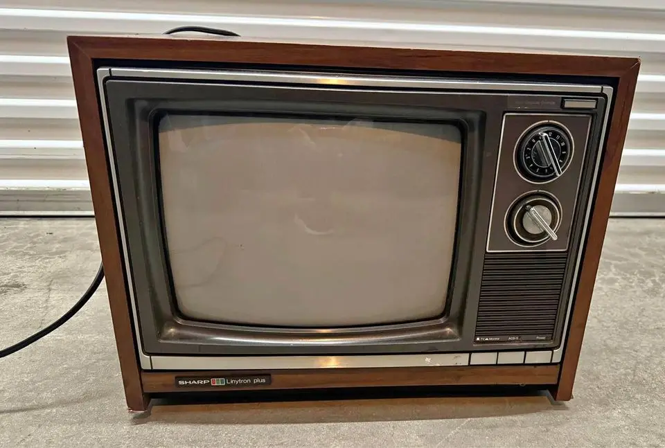 A vintage Sharp Linytron Plus television with a wooden frame and two dials sits on a concrete floor in front of a white…