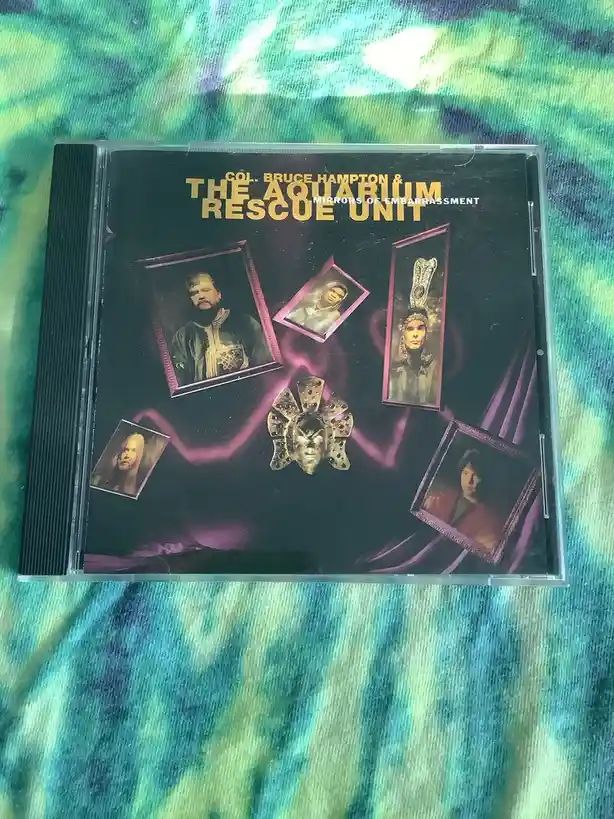 A CD case for 'Col. Bruce Hampton & The Aquarium Rescue Unit - Mirrors of Embarrassment' rests on a tie-dye fabric. The…