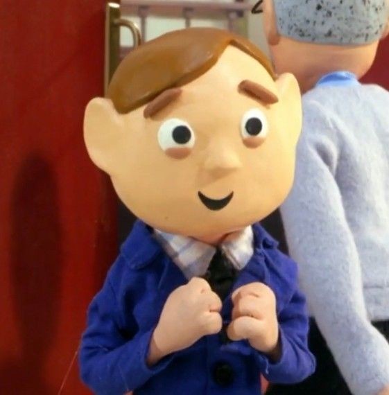 A close-up of a claymation character with large eyes and a smiling face, wearing a blue jacket and tie, standing in front of…