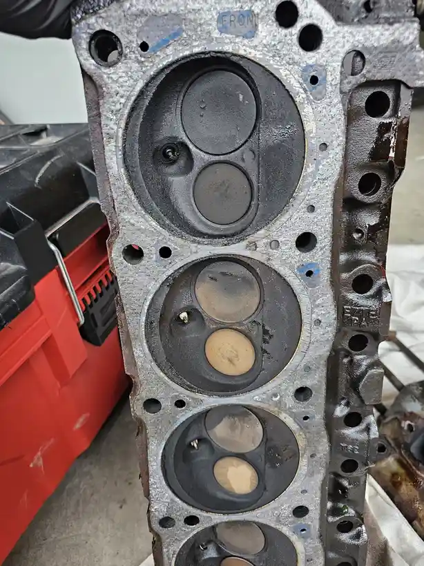 A close-up photo of a dirty, disassembled engine block with visible cylinder bores. The top cylinder has a crudely drawn sad…
