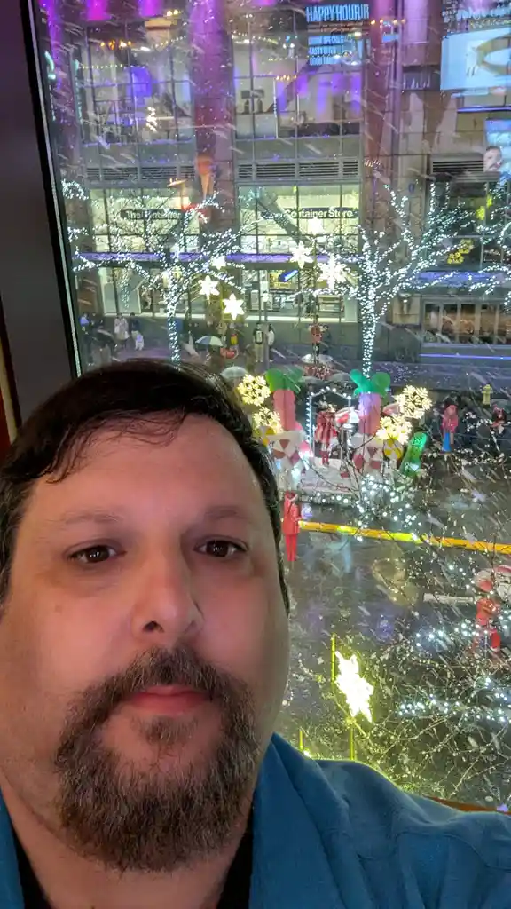 A man with a beard takes a selfie looking directly at the camera with a neutral expression, positioned indoors in front of a…