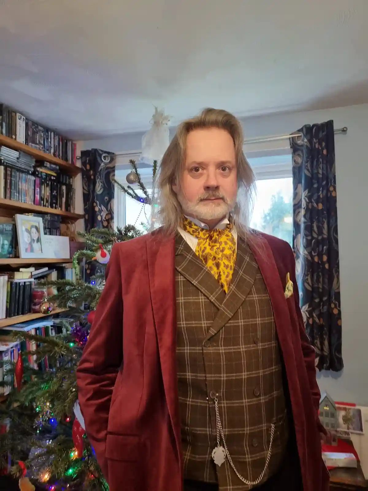 A man with long blonde hair and a beard wears a red velvet jacket over a brown plaid waistcoat and a yellow paisley cravat…