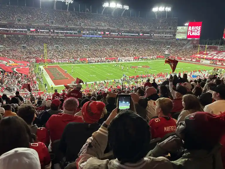 A crowded Tampa Bay Buccaneers stadium at night, filled with fans in red and gold gear, many holding flags and wearing…