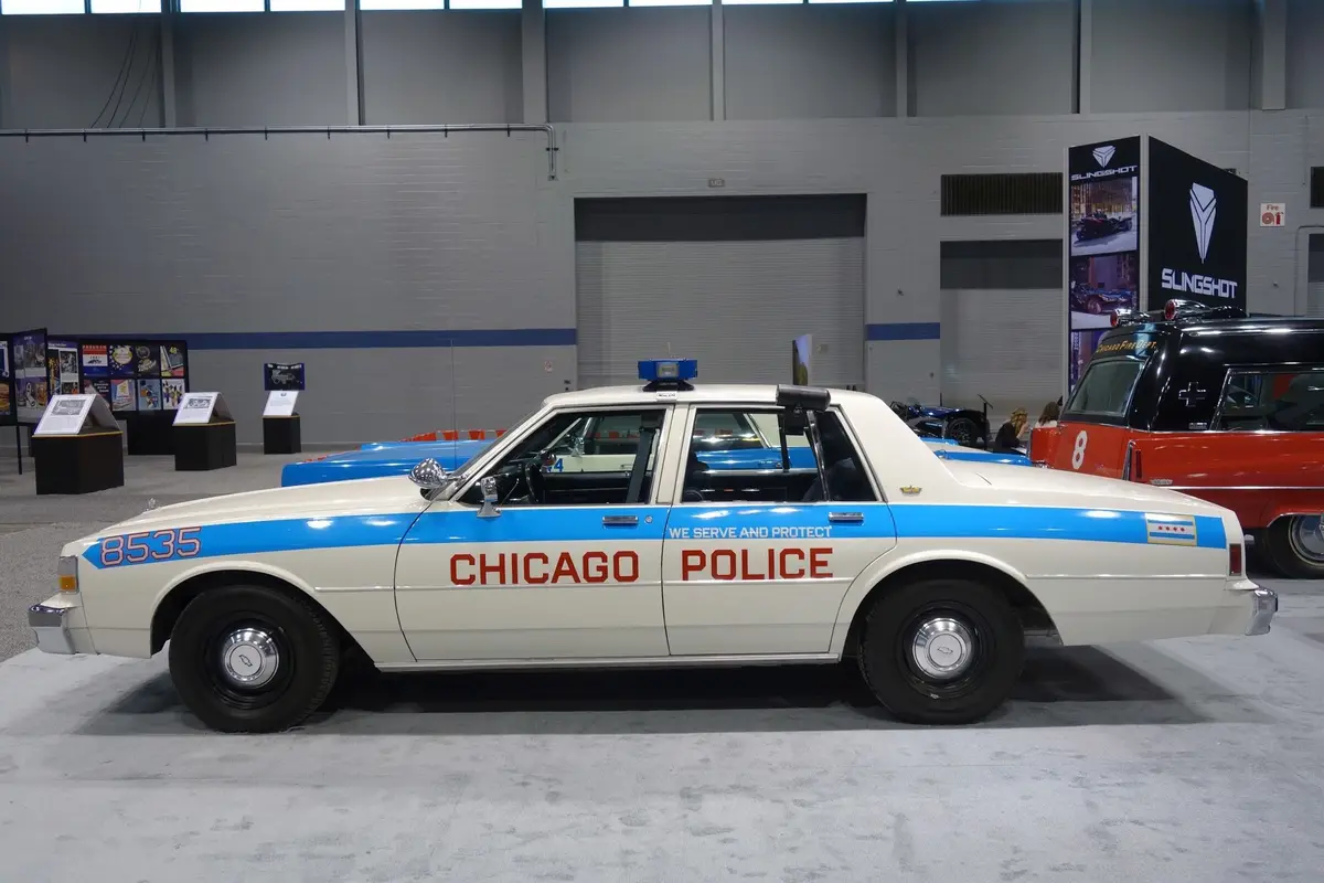 A vintage white Chicago Police car with blue stripe and red lettering is displayed indoors. The text 'WE SERVE AND PROTECT'…
