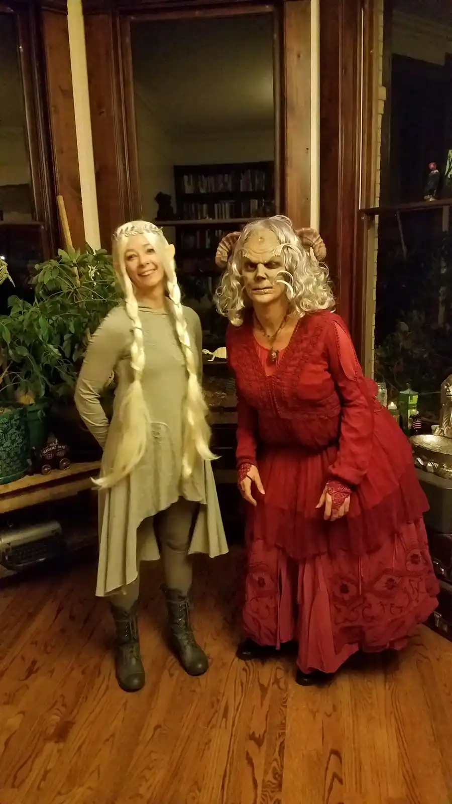 Two women in elaborate fantasy costumes stand indoors on a wooden floor. The woman on the left wears a light green tunic…