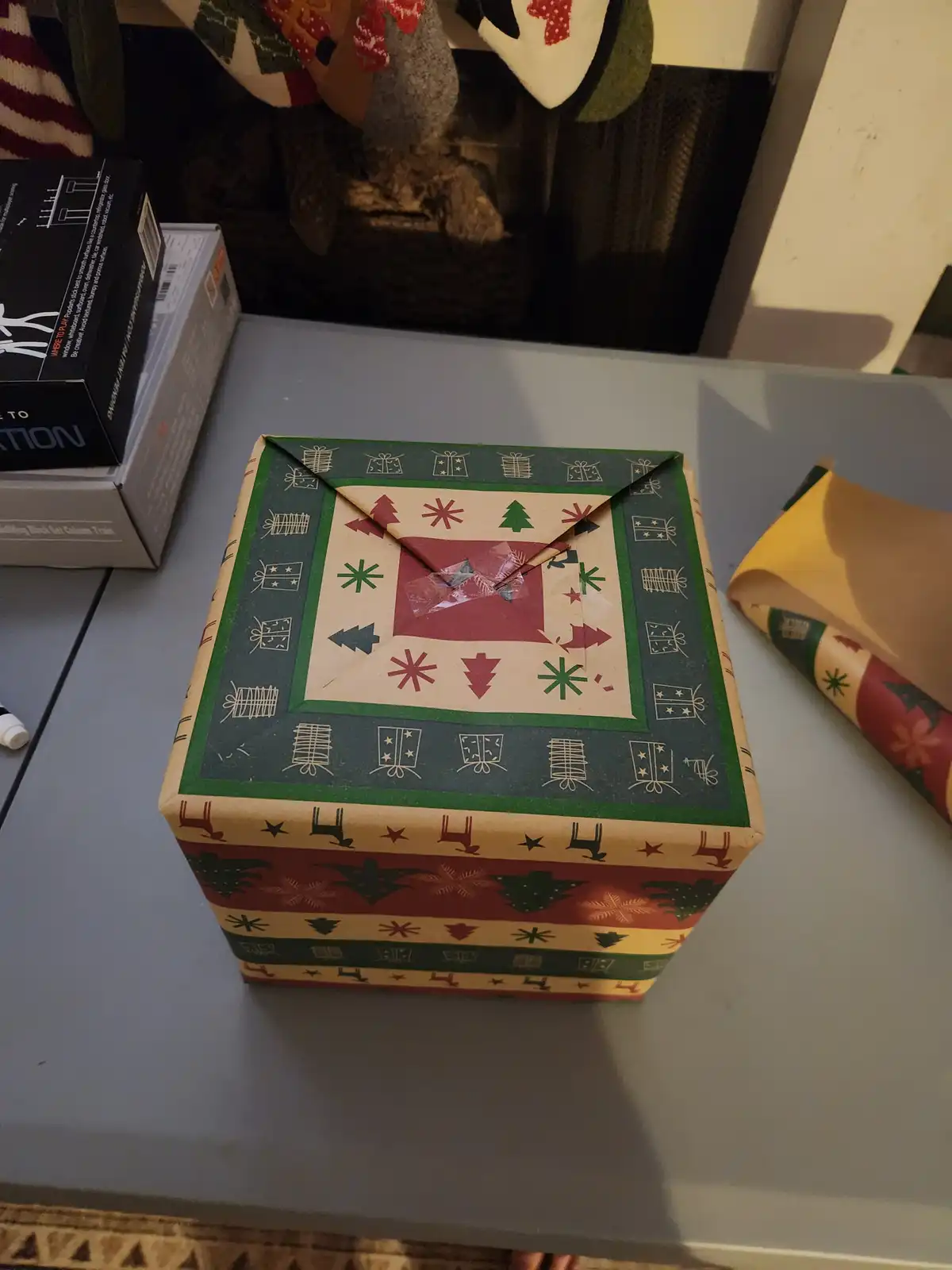 A square gift box wrapped in festive holiday paper with red, green, and beige patterns of trees, stars, and reindeer sits on…