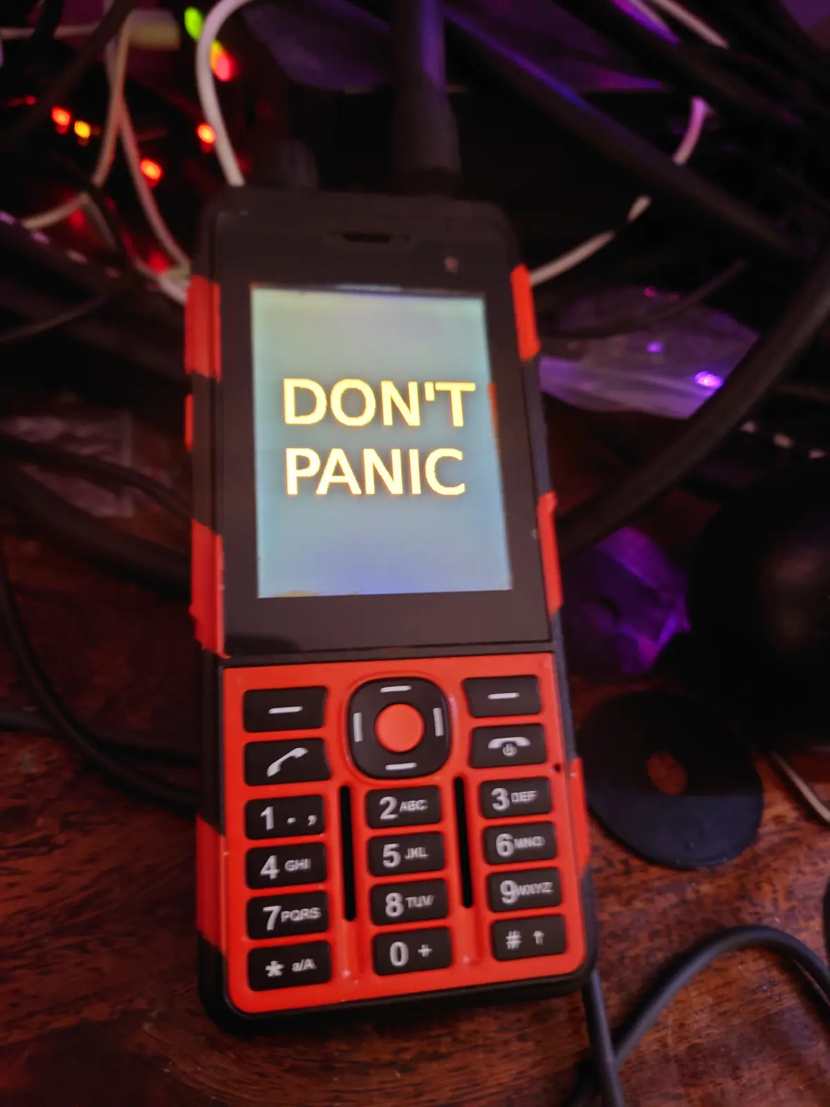 A red and black rugged feature phone with a physical keypad displays the text 'DON'T PANIC' in glowing yellow letters on its…