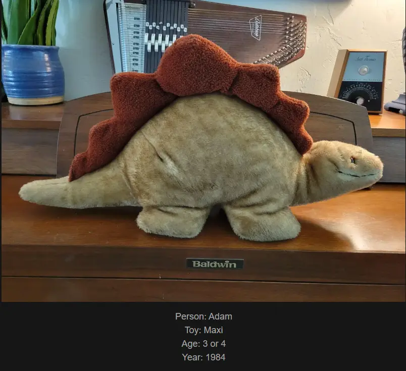 A beige plush Stegosaurus toy with a brown, fuzzy backplate sits on a wooden Baldwin piano. The toy is labeled as 'Maxi'…
