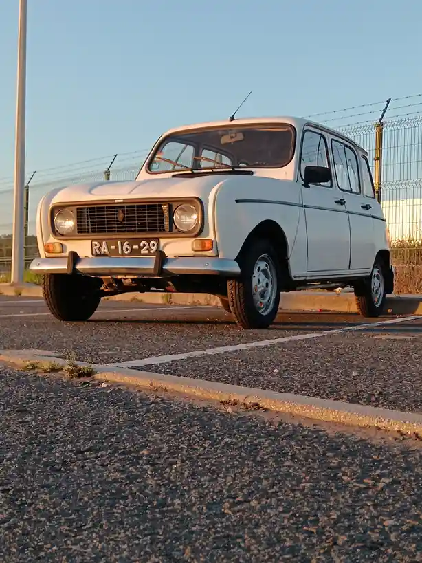 A white vintage Renault 4 parked in a lot with a fence behind it, its license plate reads 'RA-16-29', and the text 'ra 16…