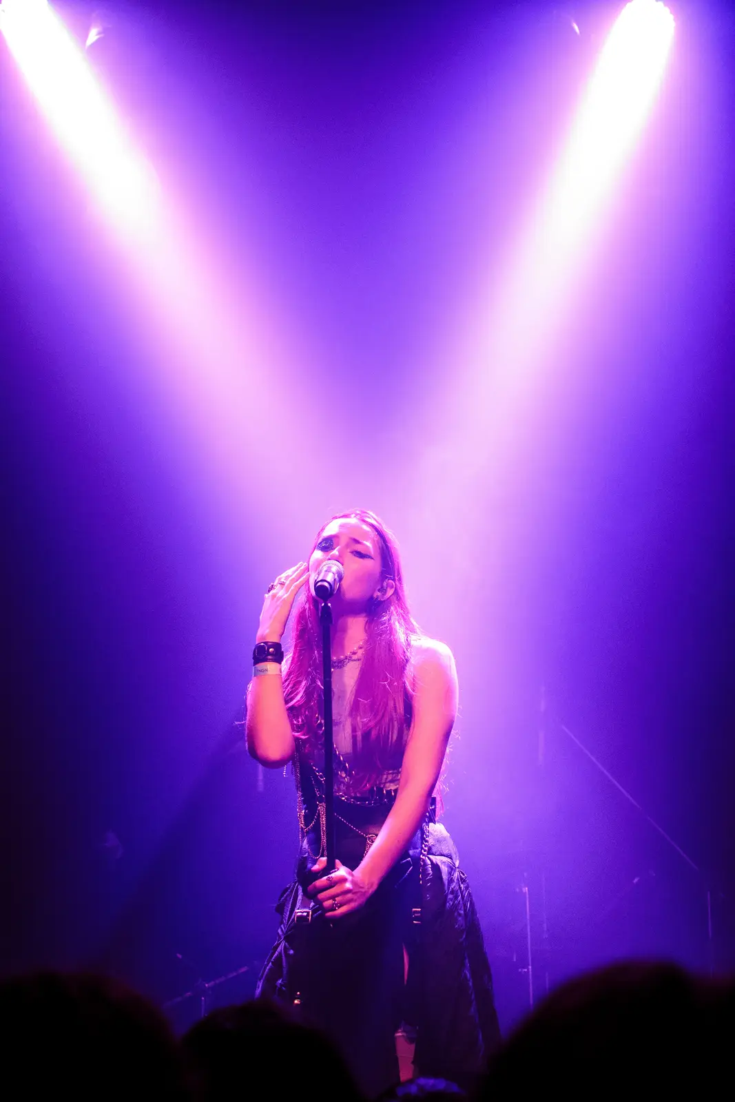 A female singer performs on stage under two bright purple spotlights, her hand near her face as if emotional. She wears dark…
