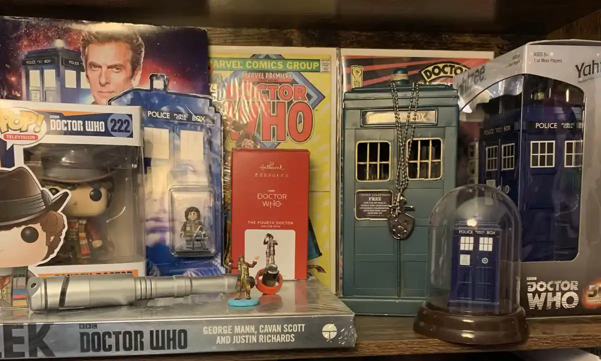 A shelf displays a collection of Doctor Who merchandise including Funko Pop! figures, LEGO sets, comic books, a Hallmark…