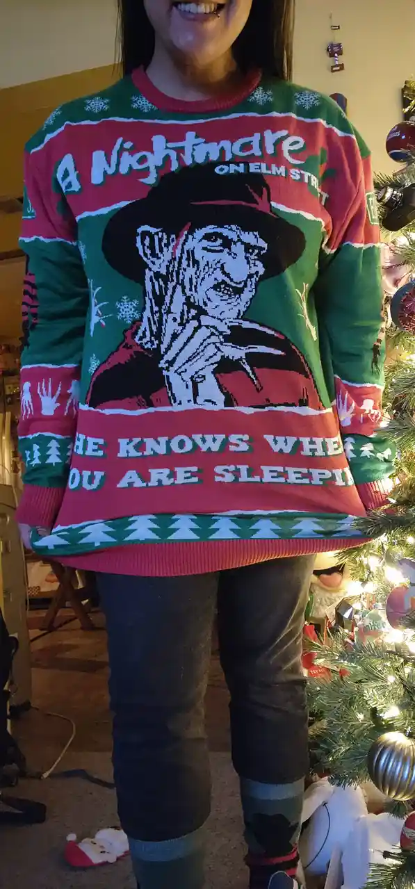 A person is smiling while wearing a red and green ugly Christmas sweater featuring the character Freddy Krueger from 'A…