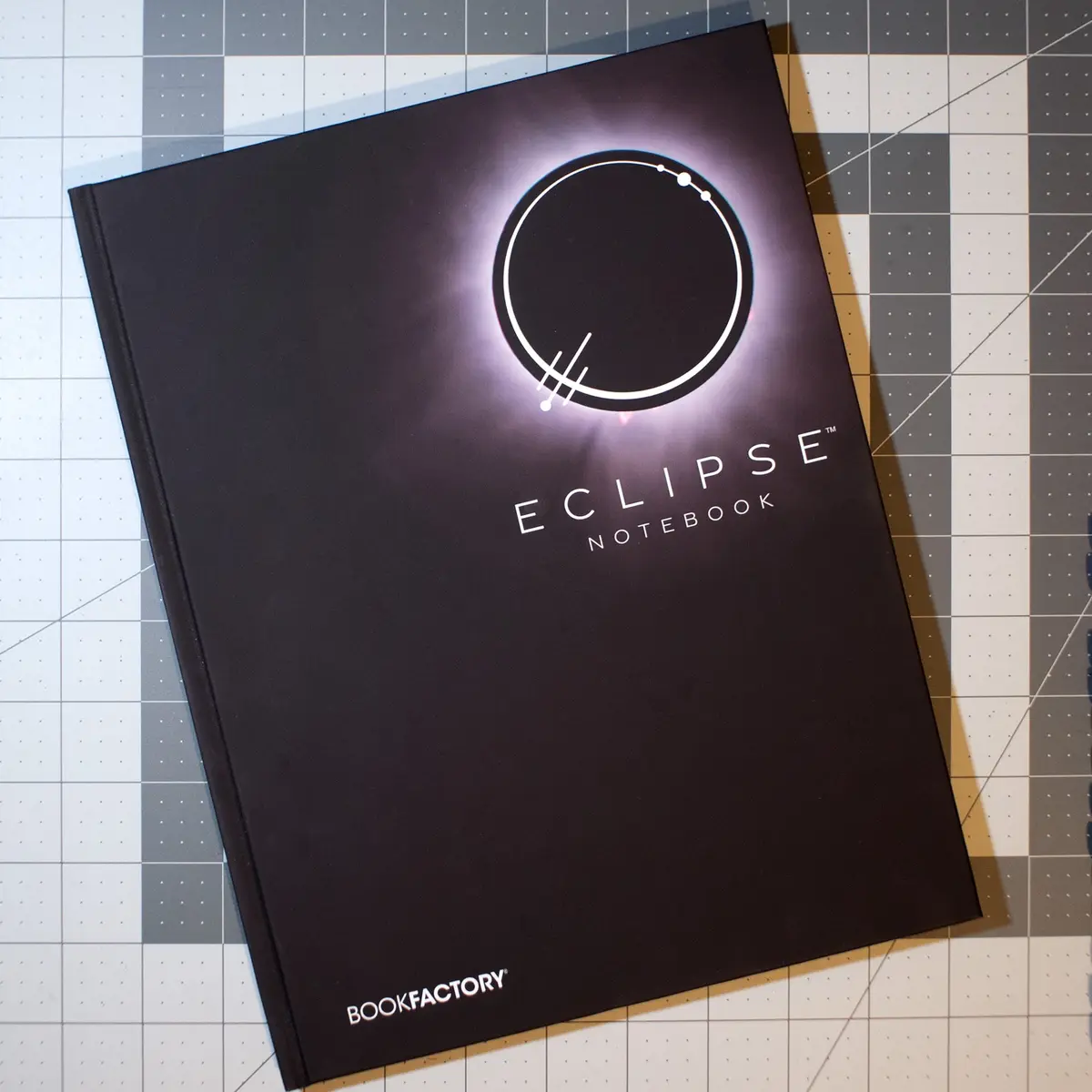 A black notebook titled 'ECLIPSE NOTEBOOK' by BOOKFACTORY rests on a gridded cutting mat. The cover features a stylized…