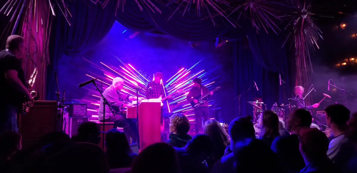 A live band performs on a dimly lit stage with purple and blue lighting, surrounded by dark curtains and starburst-like…