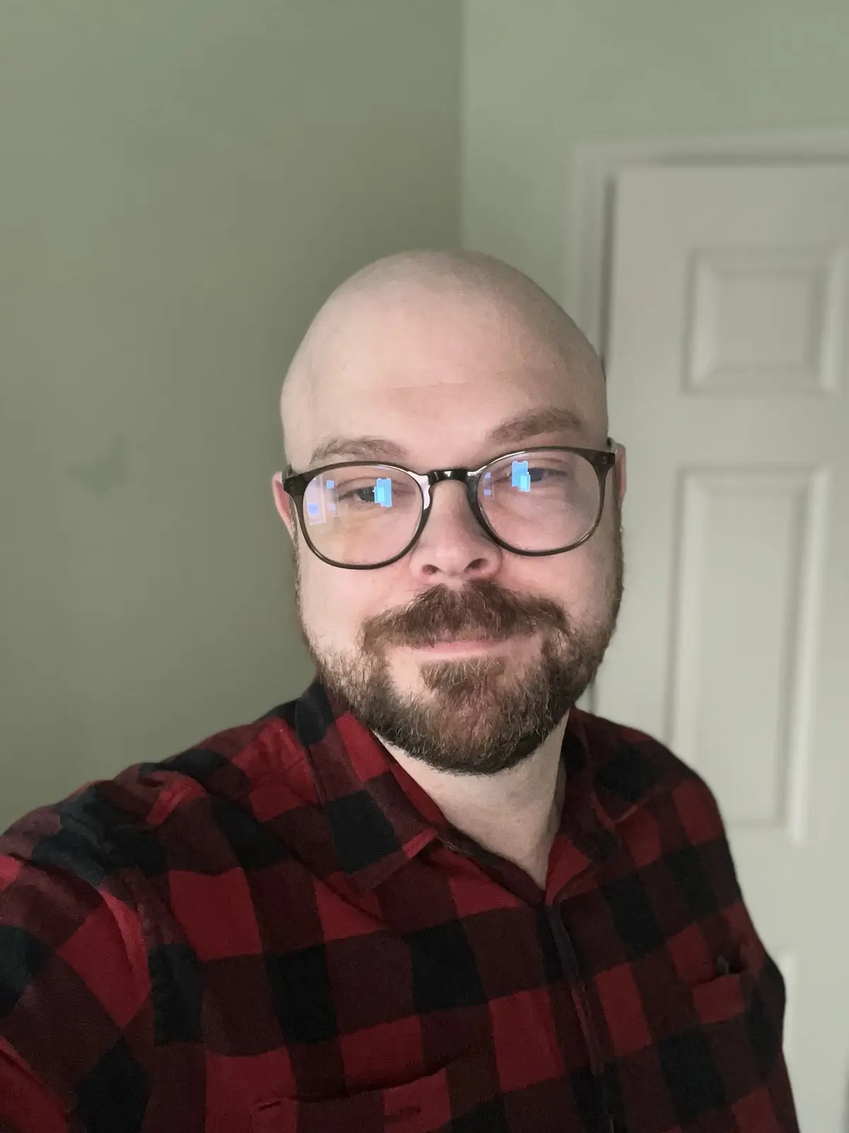 A bald man with a beard and glasses wearing a red and black plaid shirt looks at the camera with a slight smirk. The text…