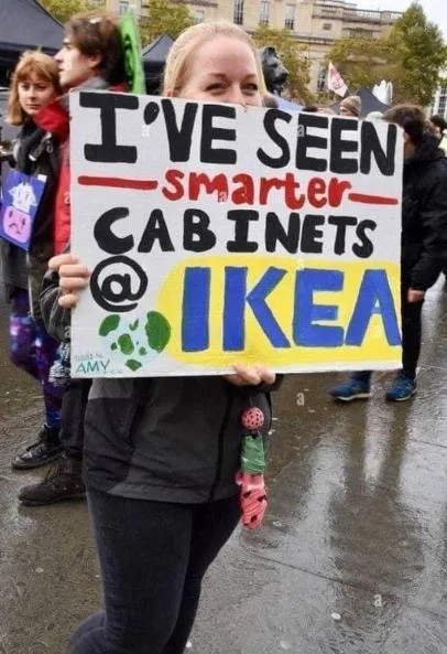 A person holds a handmade protest sign outdoors in wet weather, reading 'I'VE SEEN smarter CABINETS @ IKEA'. The sign…