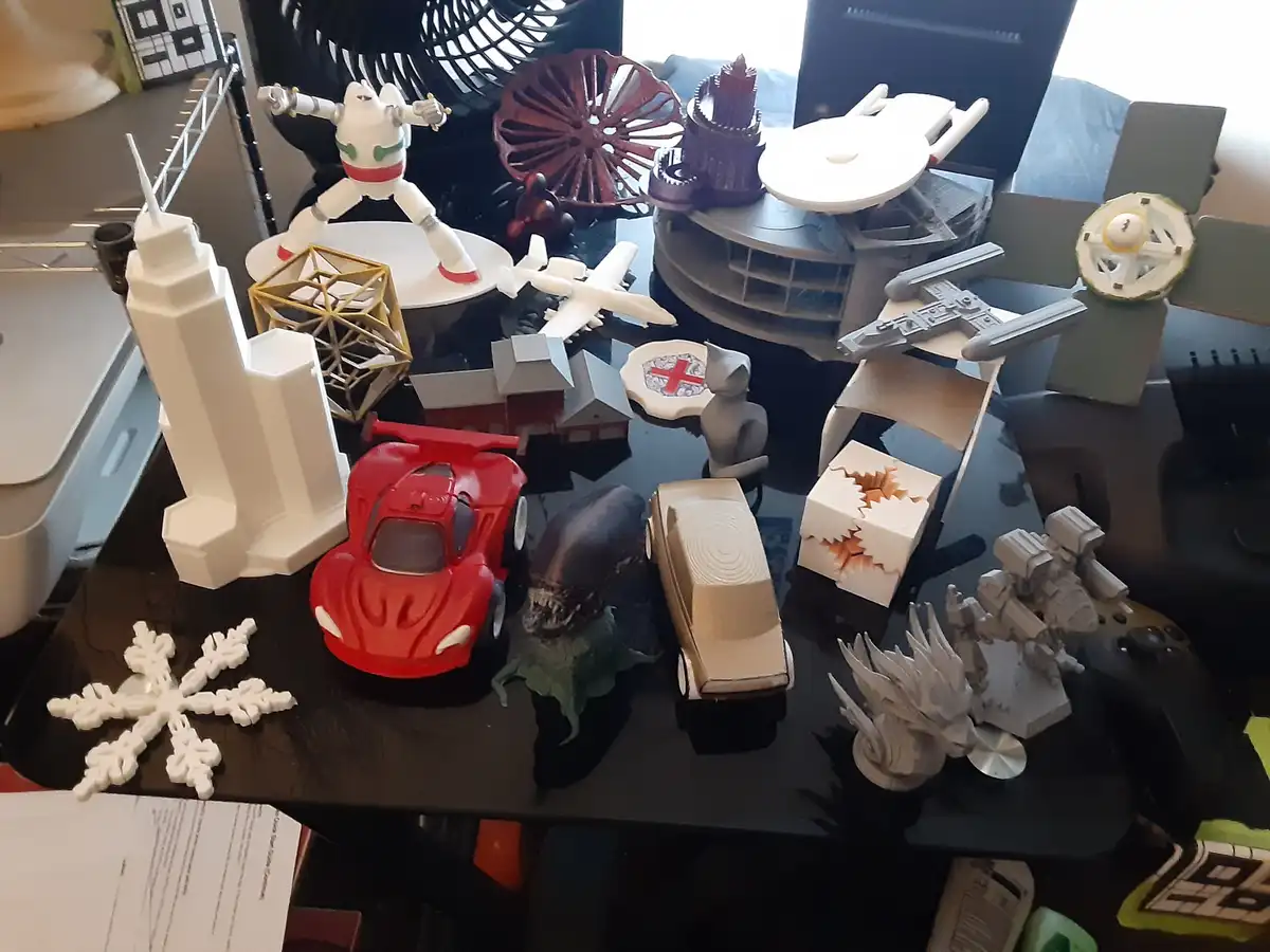 A cluttered black desk is covered with numerous 3d-printed objects including a red sports car, an alien, a white snowflake…