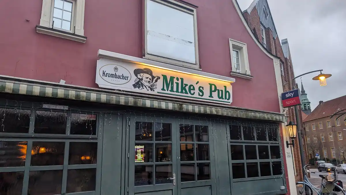 An exterior shot of a pub named 'Mike's Pub' with a lit sign featuring the Krombacher beer logo and a man's silhouette. The…