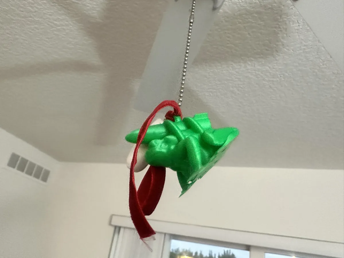 A green, textured, leaf-like ornament with a red ribbon is hanging from a silver chain attached to a white ceiling. The…