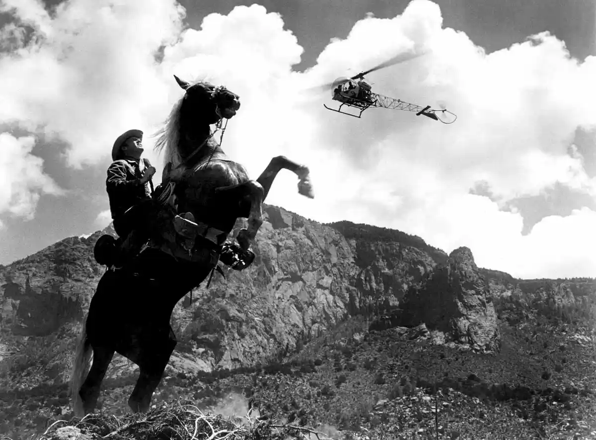 A black-and-white photo shows a man in a cowboy hat riding a rearing horse on a rocky hillside, looking up at a helicopter…