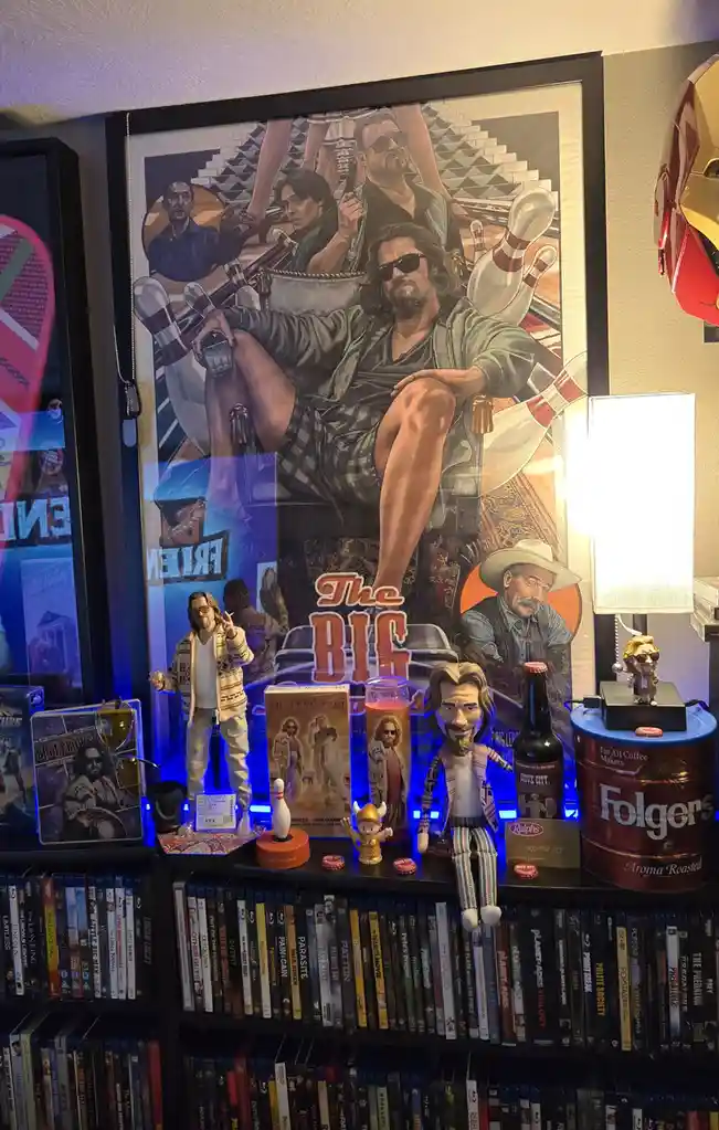 A framed poster of 'The Big Lebowski' movie is displayed above a shelf filled with movie memorabilia, including action…