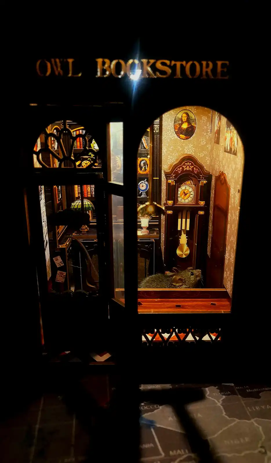 A dimly lit, ornate miniature diorama labeled 'OWL BOOKSTORE' shows a mouse sitting on a wooden bench in front of a…