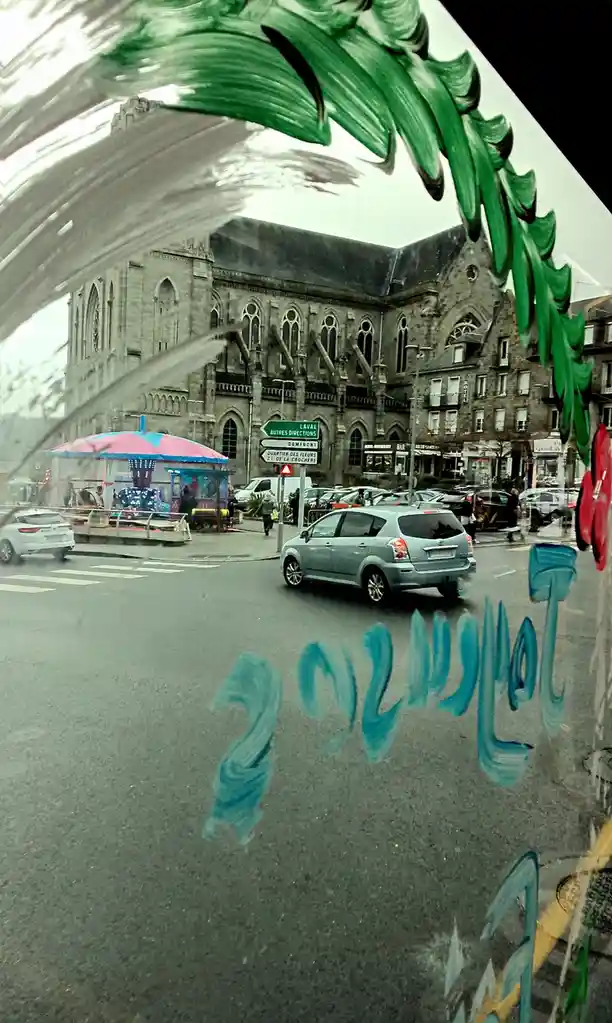 A street view through a window with green and blue graffiti, including the word 'téléphone' in blue paint, showing a…