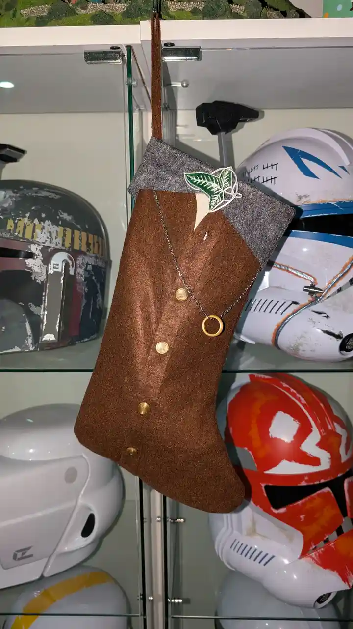 A brown felt Christmas stocking designed to look like a vest with gold buttons and a green leaf pin hangs in a glass display…