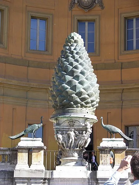 a large, ornate bronze pinecone sculpture stands on a decorative pedestal in front of a classical building with windows. Two…
