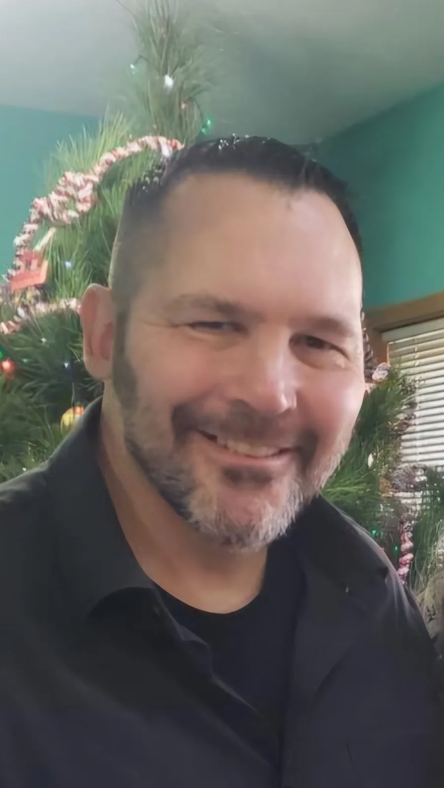 A man with a short haircut and a salt-and-pepper beard smiles at the camera while standing in front of a decorated christmas…