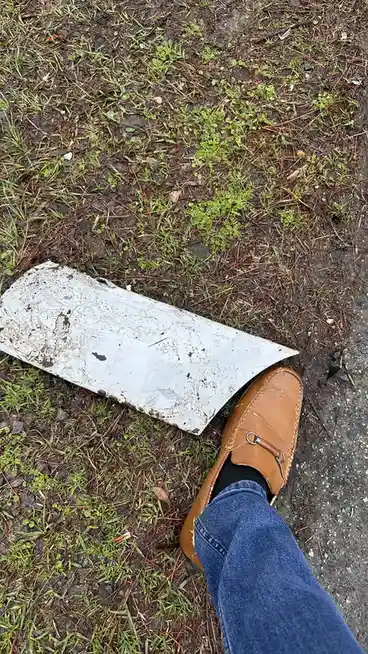 A person wearing blue jeans and a brown leather loafer stands next to a large, dirty, white triangular piece of metal lying…
