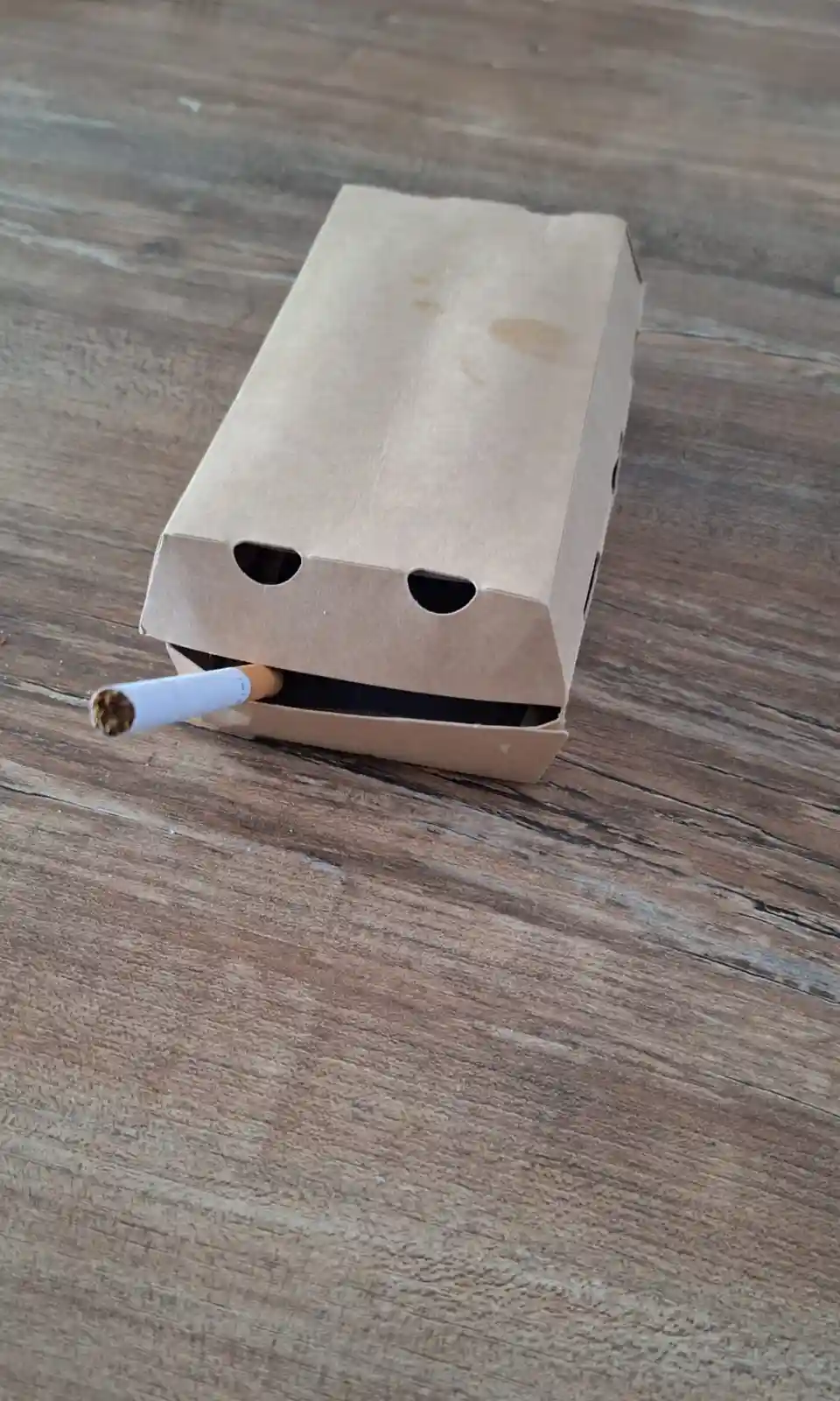 A brown paper takeout food container, resembling a face with two holes for eyes and a slot for a mouth, has a lit cigarette…