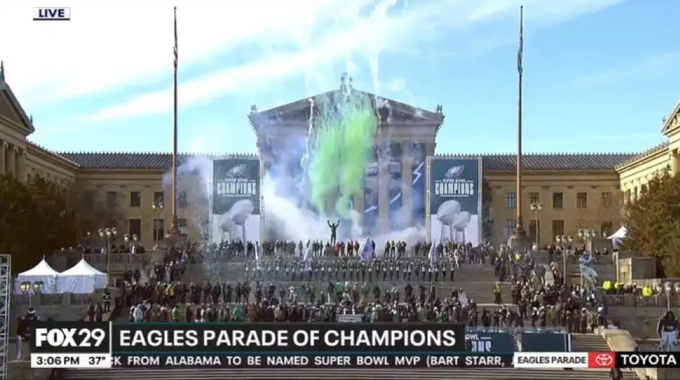 A live broadcast image shows a large crowd gathered in front of a grand classical building during a parade. Green smoke…