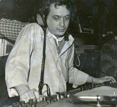 A black-and-white photo shows a man with curly hair wearing headphones and a light-colored shirt, standing behind a DJ…