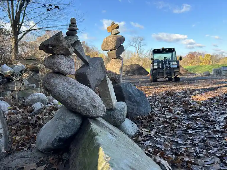 A photo shows two precarious cairns made of stacked rocks in a leaf-covered yard, with a forklift visible in the background…