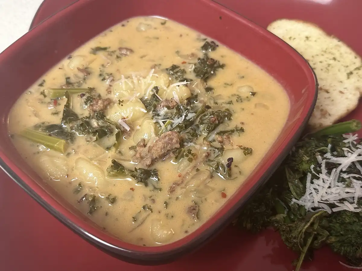 A close-up photo shows a red bowl filled with creamy gnocchi soup containing kale, sausage, and grated cheese, served with…