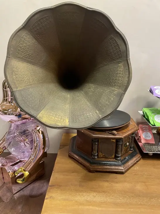 A vintage gramophone with an ornate brass horn sits on a wooden table, next to a decorative pink glass object and some…