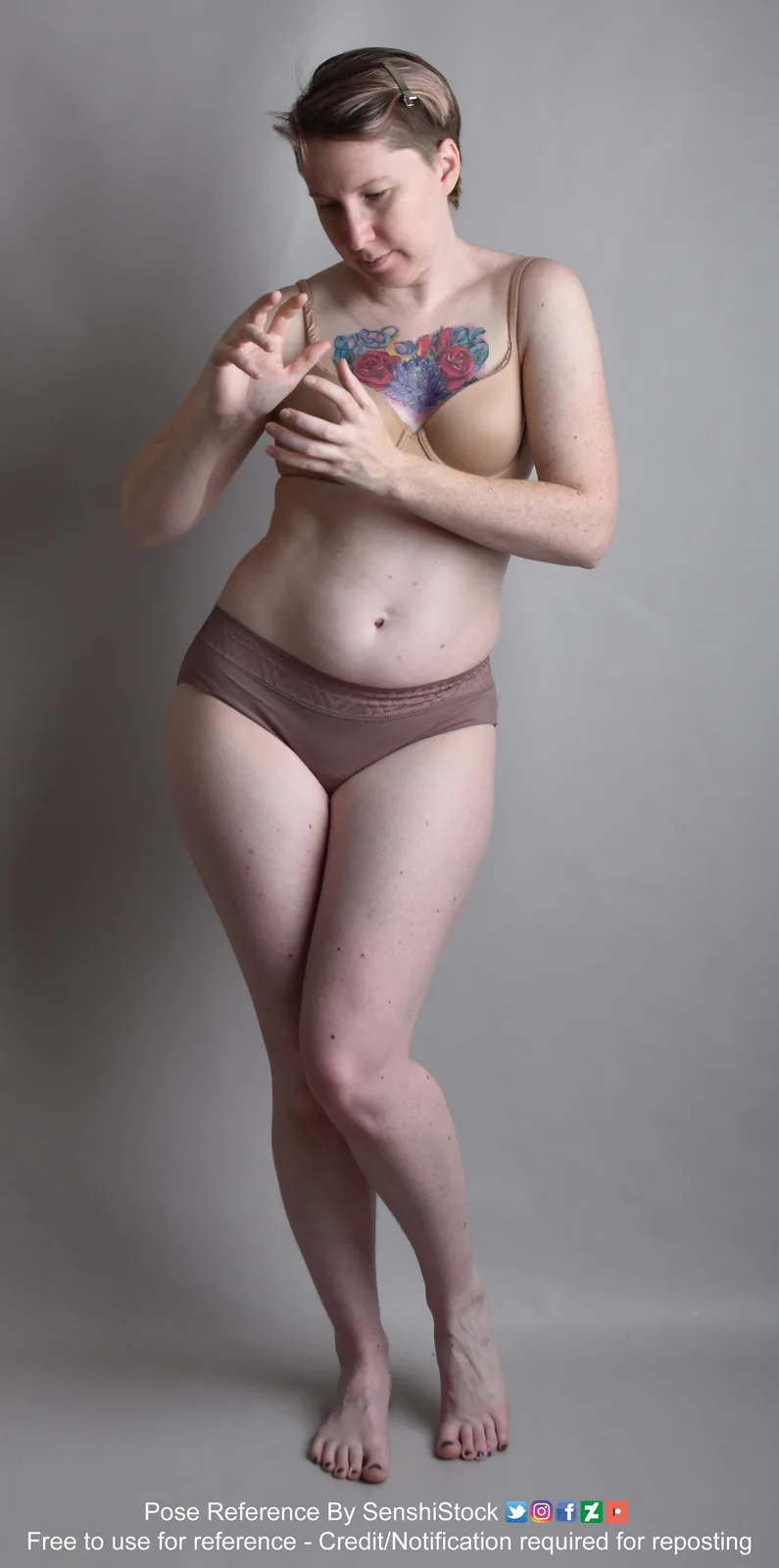 A woman with short hair and a colorful floral tattoo on her chest stands in a studio against a plain gray background…
