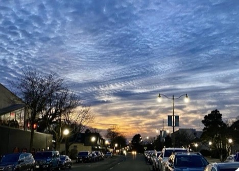 A street scene at dusk with cars parked along both sides, streetlights on, and a dramatic sky filled with textured clouds…