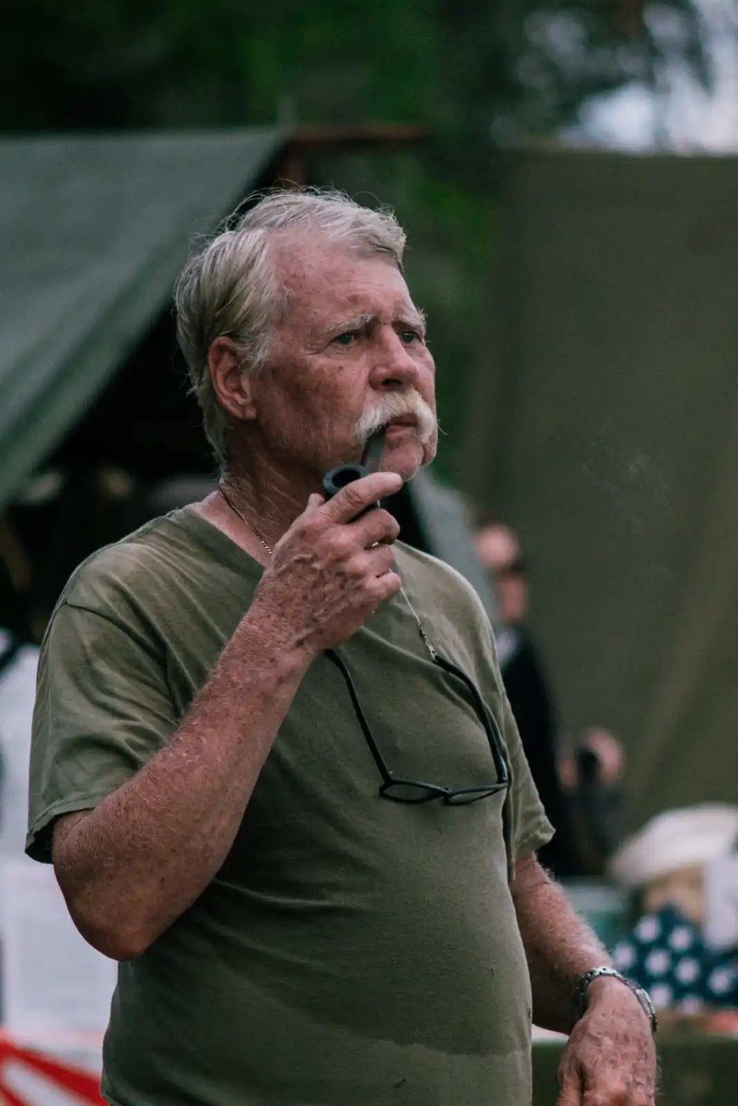 An older man with a white mustache and gray hair is seen in profile, holding a pipe to his mouth and looking off to the…