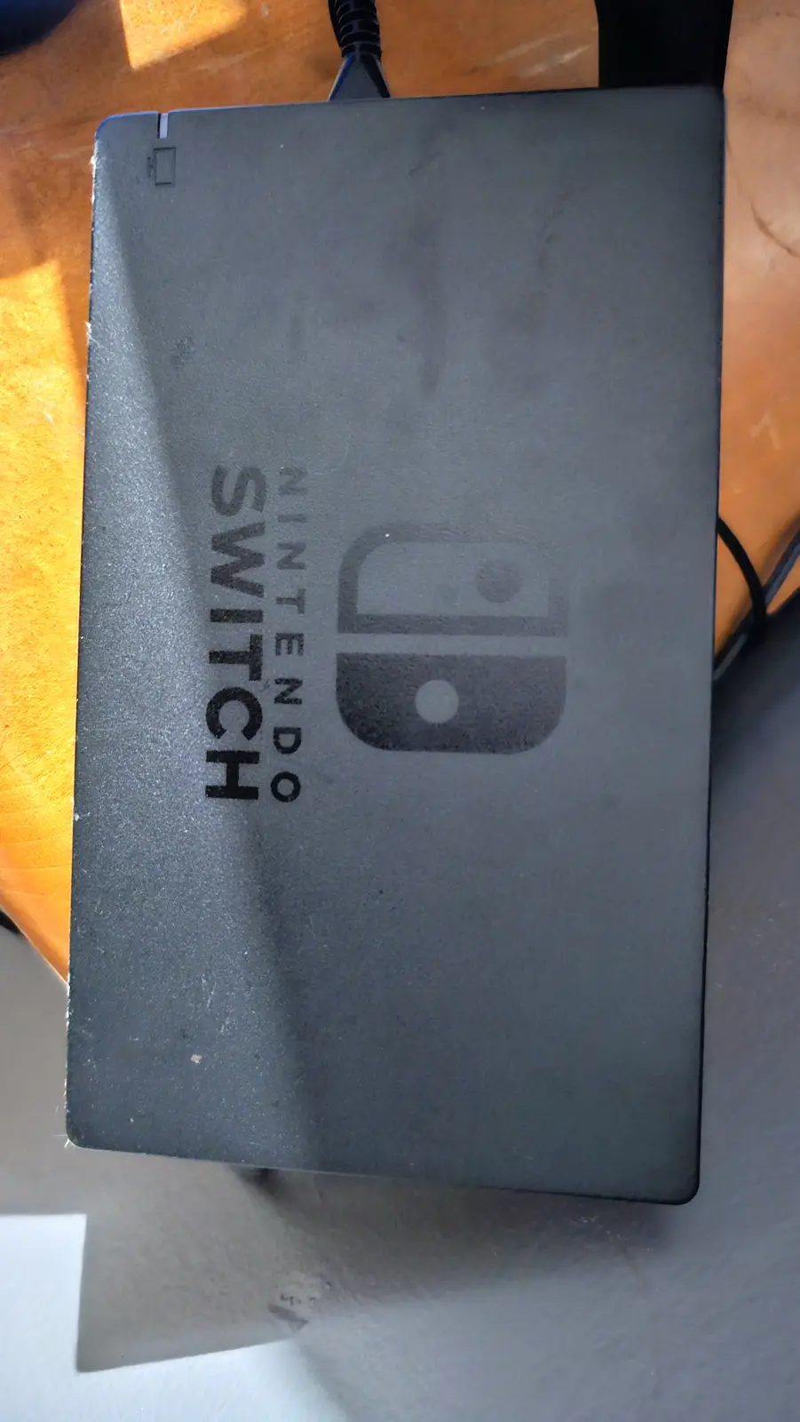 A top-down photo of a gray Nintendo Switch console resting on a wooden surface, showing its logo and text. The overlaid text…