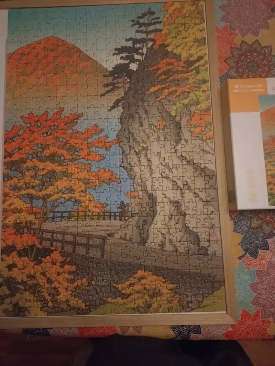 A completed 500-piece jigsaw puzzle depicting a scenic autumn landscape with vibrant orange and red foliage, a large tree, a…
