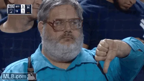 A man with a gray beard and glasses wearing a blue shirt gives a thumbs-down gesture while sitting in a stadium crowd. The…