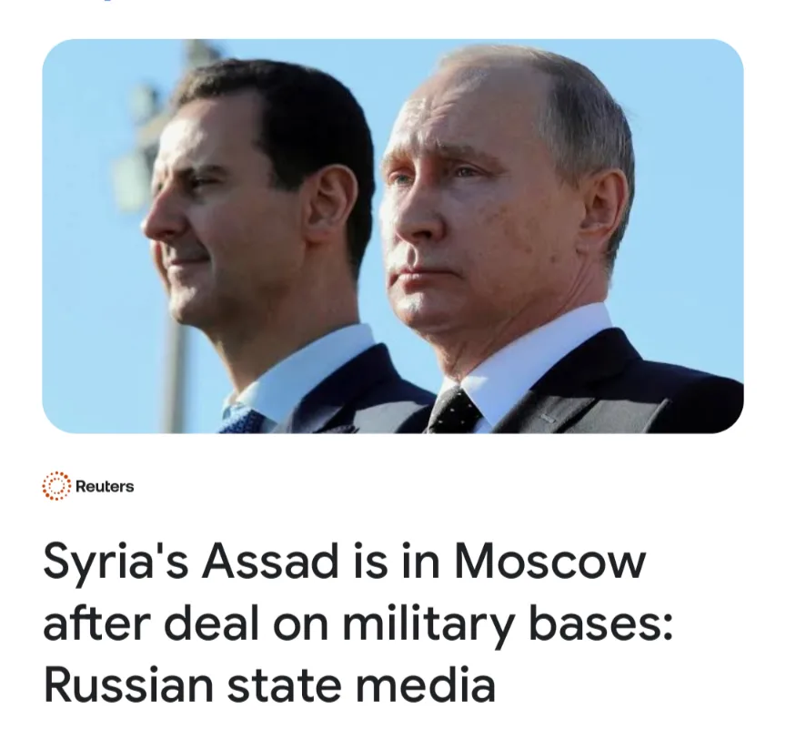 The image shows two men in suits standing side by side against a clear blue sky. The man on the left, Bashar al-Assad, has a…