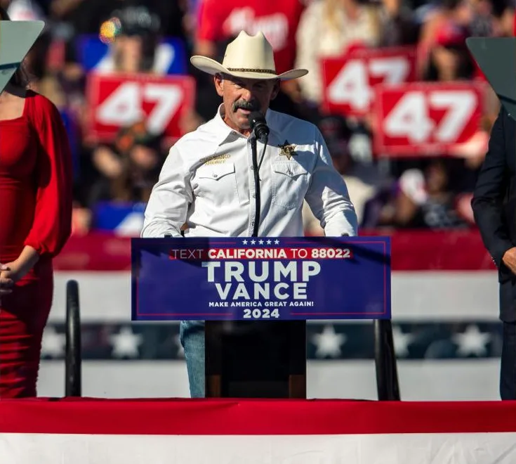 A man in a cowboy hat and white shirt stands at a podium with a blue sign that reads 'TEXT CALIFORNIA TO 88022 TRUMP VANCE…